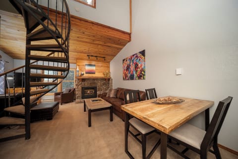 Unwind in your open-concept living space featuring a cozy stone fireplace, rustic wood dining table, and striking spiral staircase leading to your loft retreat.