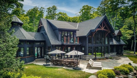 Stone patio with wooden dining set and umbrellas, flanked by cushioned lounge chairs beside dark wood cottage.