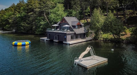Dark wood boathouse with rooftop deck, glass railing, and lakeside dock featuring a floating slide and trampoline.