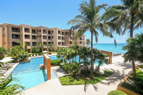 Beachfront property featuring a multi-level building, sparkling swimming pool, tropical landscaping, and direct access to white sand beach under clear blue skies.