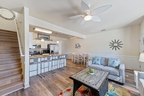 Your open-concept living space flows seamlessly from kitchen to lounge, where coastal charm meets modern comfort under gentle ceiling fan breezes.