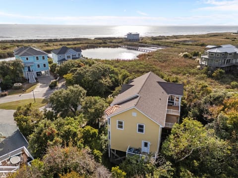 Aerial view of elevated Oak Island beach home surrounded by coastal greenery, nearby homes, scenic pond, and easy access to the ocean just beyond the dunes. Perfect location for a relaxing beach getaway.