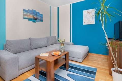 A cozy living area with a gray sofa, a small wooden coffee table, and a vase with green plants. The walls are painted in bright blue, and a decorative painting is mounted on the wall.