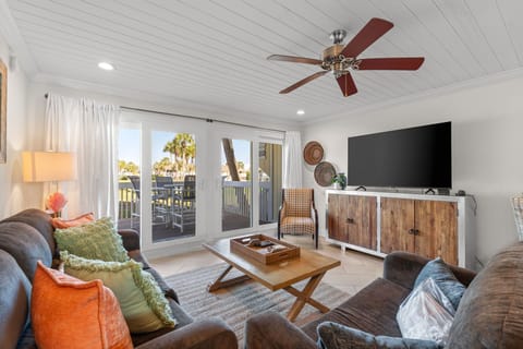 Your cozy living room invites you to unwind on plush seating beneath a gentle ceiling fan, with French doors opening to your private balcony and tropical views beyond.