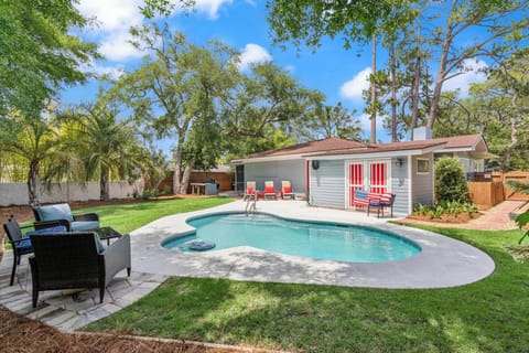 A private pool with spa awaits in your lush, tree-shaded backyard, complete with lounge seating, red Adirondack chairs, and a cheerful poolside cottage for storage or relaxation.