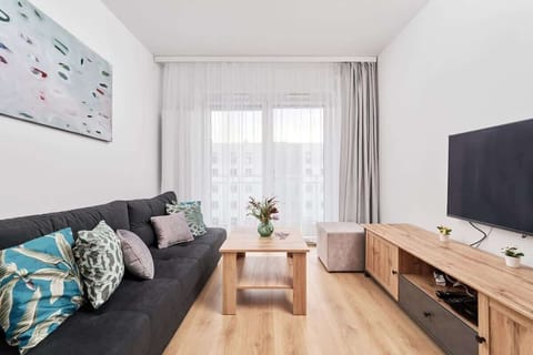 A living room featuring a gray sofa, a wooden coffee table, and modern decor. Large windows allow natural light to fill the space.