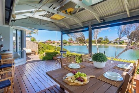 This beautiful covered deck overlooks a serene waterfront, offering a perfect setting for outdoor dining and relaxation. A wooden table set for four with plates and a cheese platter creates an inviting atmosphere, while the surrounding greenery and water 