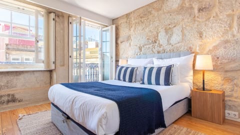 Drift off to sleep in the plush double bed, surrounded by the inviting charm of exposed stone walls and bathed in natural light from the large windows. The open balcony doors invite a gentle breeze, creating a perfect sanctuary for rest and rejuvenation. 