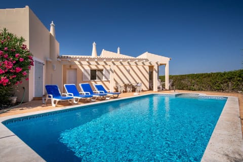 This sunny image reveals a beautiful outdoor area featuring an inviting blue pool, perfect for swimming and relaxation. In the background, a charming light-yellow holiday home exudes a cozy atmosphere. Comfortable blue sun loungers around the pool beckon guests to soak up the warm sunshine. A vibrant pink flowering bush adds a cheerful splash of color, and the bright blue sky promises a perfect vacation day.