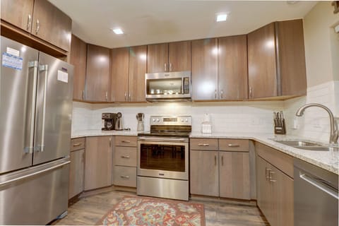 A modern kitchen with stainless steel appliances, including a refrigerator, microwave, and stove, brown wooden cabinets, a coffee maker, white subway tile backsplash, granite countertops, and a patterned rug.