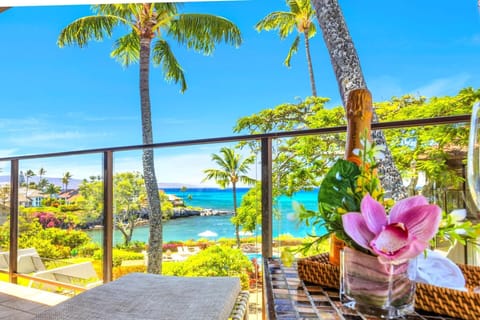 Stunning ocean-view lanai with lush tropical scenery, swaying palms, and a sparkling blue bay — perfect for enjoying a glass of champagne in paradise.