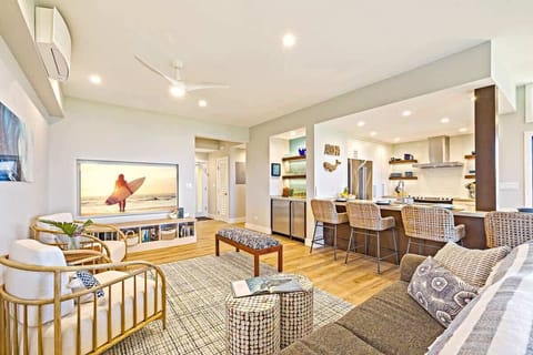 Spacious and stylish living area featuring a huge smart flat screen TV, cozy sectional sofa, and modern coastal décor. Open-concept design flows seamlessly into the kitchen with bar seating, perfect for family gatherings and entertaining in comfort.