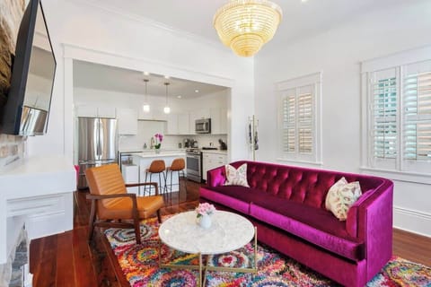 Stylish living space with a bold velvet sofa, vibrant rug, and elegant chandelier. Open layout flows into a bright, modern kitchen, creating a perfect spot to relax or gather.