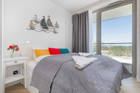 A cozy bedroom featuring a double bed with neutral bedding, decorative pillows, and nautical-themed artwork on the wall. Large windows allow plenty of natural light.