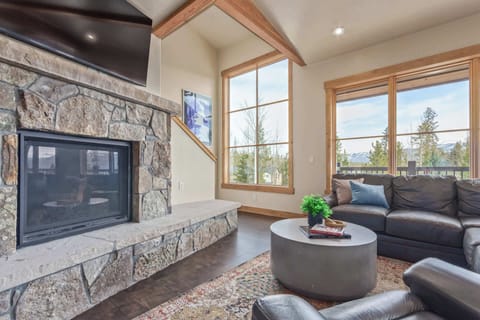 The spacious living area is anchored by a stunning stone hearth and fireplace with a large, flat-screen smart TV and bathed in natural light from the surrounding windows, offering tranquil nature views.