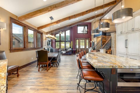Spacious open-concept kitchen and living room with vaulted ceilings, exposed wooden beams, granite countertops, large windows, and modern light fixtures.