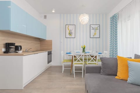 A well-lit, open-plan kitchen connected to a dining space with a wooden table and colorful accents. The bright interior feels fresh and modern.
