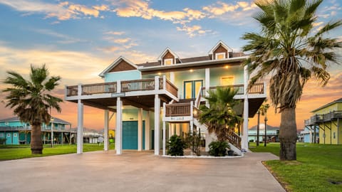 Elevated Crystal Beach vacation home with palm landscaping, spacious parking, covered ground-level space, multiple decks, and warm sunset lighting.