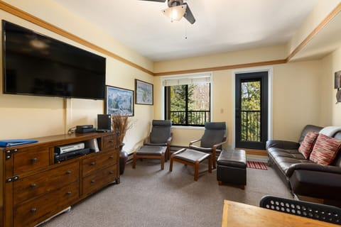 Living room with a mounted flat-screen TV, two lounge chairs with footrests, a black leather sofa, a wooden chest of drawers, and a balcony with a view of trees.