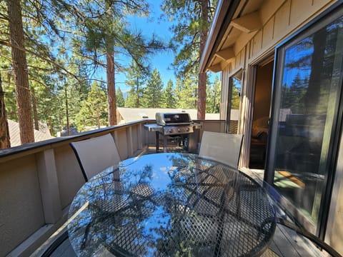 Deck with BBQ grill, patio table, patio chairs, railing, trees, rooftops, and mountain view.