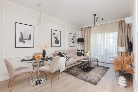 A well-lit living room with a beige sofa, decorative pillows, a glass coffee table with flowers, and modern wall art. The room is bright and styled with contemporary decor.