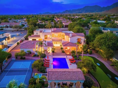 Aerial view of a stunning Mediterranean-style estate nestled in a lush desert neighborhood, featuring a glowing pool, tennis court, manicured gardens, and dramatic mountain backdrop at twilight.