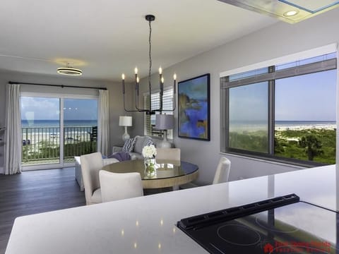 Glass-top dining table surrounded by beige chairs sits beside a window revealing ocean views.