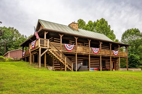 Two-level log cabin features a green metal roof, stone chimney, and a wraparound wooden deck with patriotic bunting.