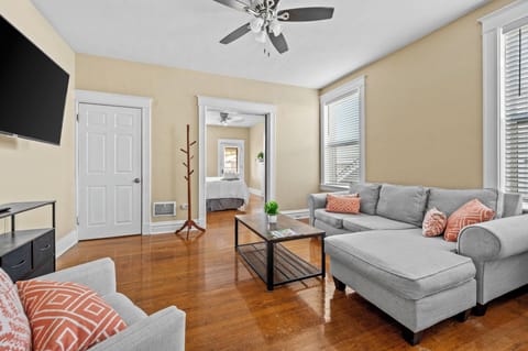 The bright living room features a gray chaise sectional, matching armchair, orange-patterned pillows, a wood-and-metal coffee table, wall-mounted TV, hardwood floors, and ceiling fan.