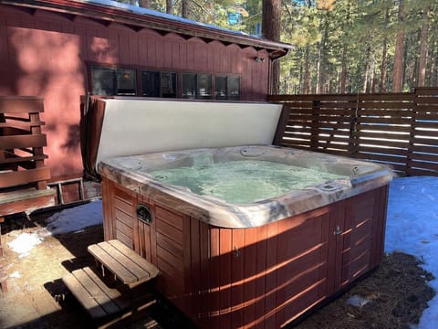 Hot tub area with open cover, steps, privacy fence, house exterior, windows, snow, and trees.