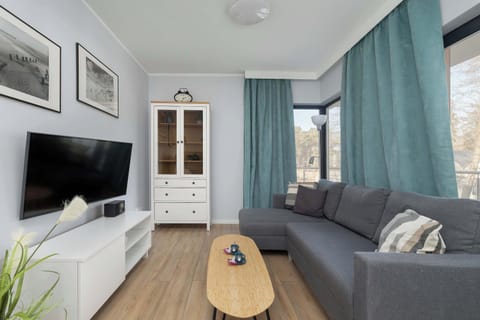 A living room with a gray couch, a wooden coffee table, and a TV. Large windows with curtains let in natural light. The space feels open and comfortable.