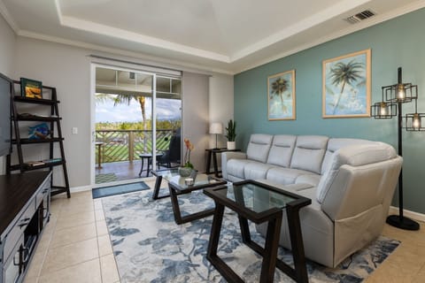 - Living room with plush sectional sofa glass coffee tables & tropical wall art accents - Sliding door opens to covered balcony with seating & views of palm lined landscape - Soft area rug and calming color palette create a relaxed welcoming atmosphere