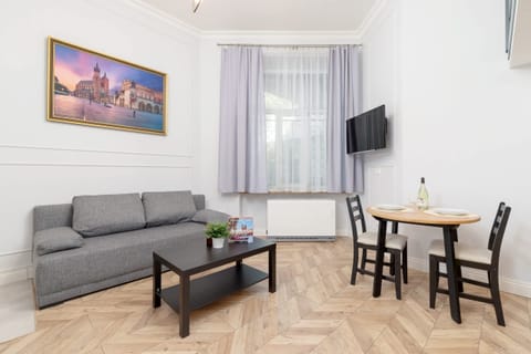Living rooing room with a gray sofa, a small black coffee table, and a table with two chairs. A wall-mounted TV and framed artwork are visible.m 