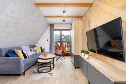 A cozy living room with a wooden dining table, chairs, and a sofa. Large windows let in natural light and offer a bright atmosphere.