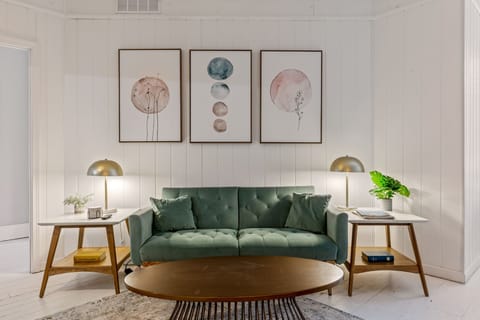 Centered living room view with a tufted green sofa, oval wood coffee table, matching side tables, brass lamps and three framed prints.