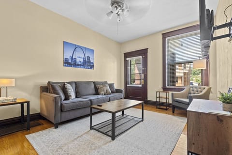 The living room features a gray sofa, tufted armchair, wood coffee table, wall-mounted TV, pale area rug, hardwood floors, large window, and windowed door.