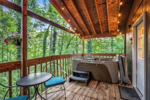 Unwind in the private covered deck featuring a hot tub, bistro seating, and string lights surrounded by peaceful forest views.