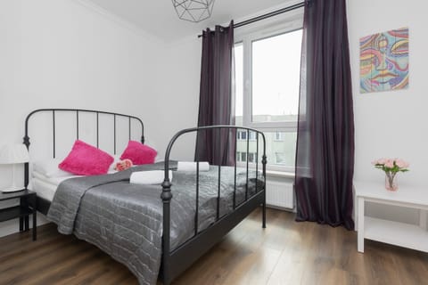 A cozy bedroom featuring a metal-framed bed, pink and gray accents, and large windows with curtains for natural light.
