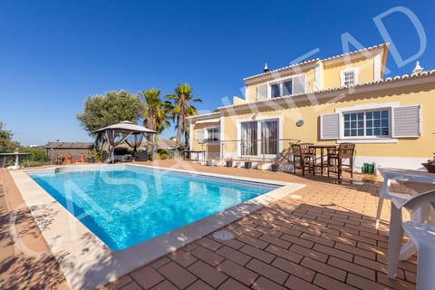 Inviting swimming pool at Casa Sinead, a spacious 3-bedroom holiday villa in the Algarve, featuring a large sun terrace with loungers and parasol beside the crystal-clear pool, with the charming villa and lush palm trees in the background