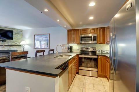 A modern kitchen with stainless steel appliances, brown cabinetry, a black countertop, a dishwasher, and a double sink. A dining area with high stools and a living room are visible in the background.