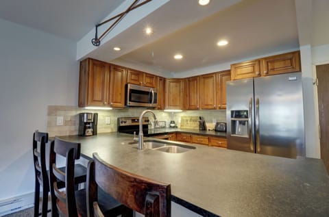 Modern kitchen with wooden cabinets, stainless steel appliances, and a central island featuring a black countertop, sink, and four high-back chairs. Under-cabinet lighting illuminates the workspace.