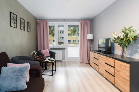 A cozy living room with a dark brown sofa, a light-colored wooden TV console, and a window with pink curtains allowing natural light.

