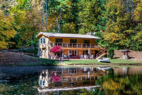 Two-story cabin with stone lower level, wooden upper deck, patio furniture, and autumn trees reflected in the lake.