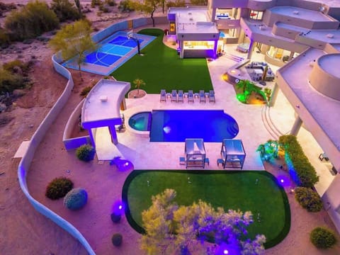 An aerial view showcases the modern luxury property set against desert landscapes, featuring a private pool, basketball court, manicured lawns, and contemporary architecture with distinctive lighting.