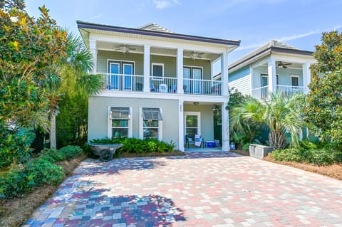 Elegant coastal home with covered porch, tropical landscaping, and decorative brick driveway creates a welcoming arrival experience.
