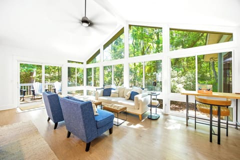 Vaulted ceilings and floor to ceiling windows frame a leafy forest backdrop, filling this airy lounge with a soft afternoon glow. Indigo armchairs and warm wood textures create a bright, calm spot to gather or read.