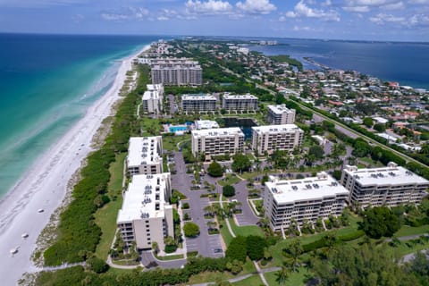 Aerial view of beachfront condos with white rooftops, green lawns, palm trees, and the sandy shoreline along turquoise waters.