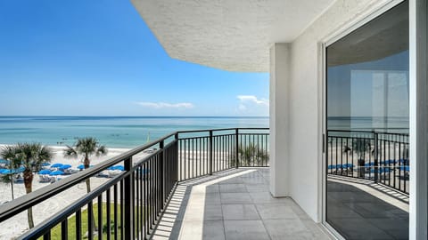 Tiled balcony with black railing overlooks white sandy beach, palm trees, and calm ocean waters under clear blue sky.