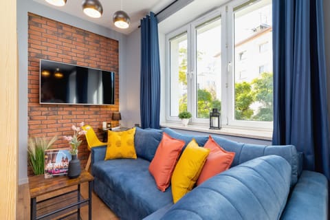 A cozy living room with a blue sofa, colorful pillows, and a brick accent wall, creating a modern and inviting ambiance.
