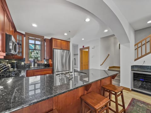 Modern kitchen featuring stainless steel appliances, dark granite countertops, wooden cabinetry, and a center island with bar stools. Stairs are visible leading to another level of the home.